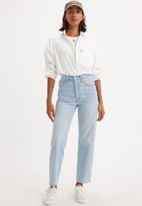 Jeans claros de cintura alta combinados com uma camisa branca de botão e um boné cinza; apresenta ténis e acessórios de joalharia discretos.