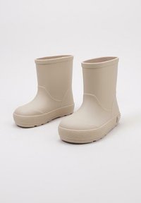 Bottes en caoutchouc beige avec une finition lisse, bout rond, design à cheville basse, semelle texturée et branding discret sur le côté.