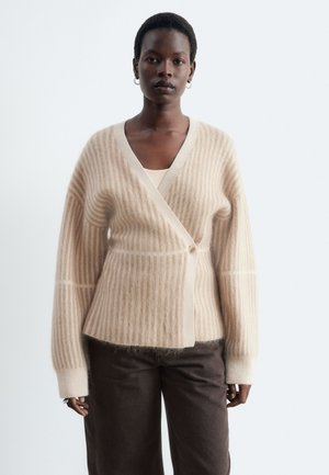 Femme aux cheveux courts portant des boucles d'oreilles créoles, un cardigan beige rayé avec un bouton, et un pantalon marron foncé, debout devant un fond uni.
