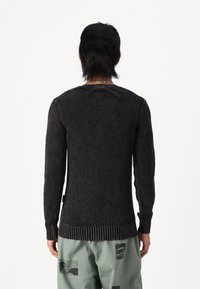 Homme portant un pull texturé noir et un pantalon gris clair avec des taches noires abstraites, debout de dos sur un fond blanc.