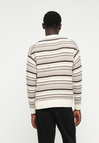 Pull en tricot avec un motif rayé crème et marron, ourlets et poignets côtelés, encolure ronde et finition texturée.