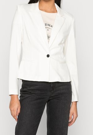 Kvinde iført en tætsiddende hvid blazer med en enkelt sort knap over en grafisk t-shirt og sorte jeans, stående mod en ensfarvet baggrund.