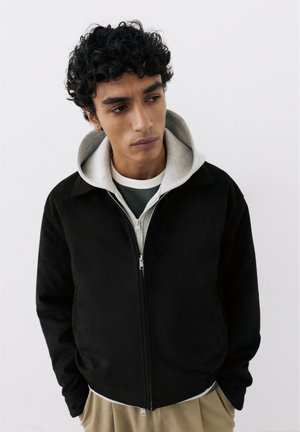 Jeune homme aux cheveux bouclés et foncés portant une veste noire zippée par-dessus un sweat à capuche gris et un pantalon beige, regardant vers le bas et sur le côté.