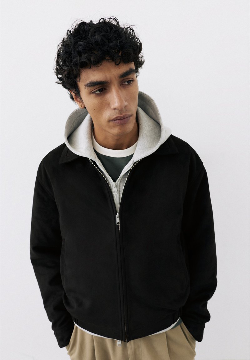 Jeune homme aux cheveux bouclés et foncés portant une veste noire zippée par-dessus un sweat à capuche gris et un pantalon beige, regardant vers le bas et sur le côté.
