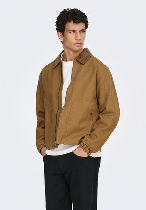 Giovane uomo con capelli ricci che indossa una giacca marrone con cerniera e una camicia bianca, con le mani nelle tasche, guarda verso destra su uno sfondo semplice.