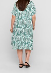 Vihreä ja valkoinen kuvioitu mekko, jossa on lyhyet hihat, kerroksellinen hame ja rento istuvuus. Yhdistetty mustiin sandaaleihin, jossa näkyy tatuointi jalassa.