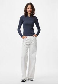 Jovem mulher com cabelo encaracolado, vestindo uma blusa de manga longa azul marinho, calças brancas de perna larga e ténis prateados, de pé com as mãos nos bolsos.