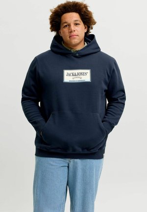 Jong persoon met krullend haar die een marineblauwe Jack & Jones-hoodie en lichtblauwe spijkerbroek draagt en met de handen in de zakken staat.