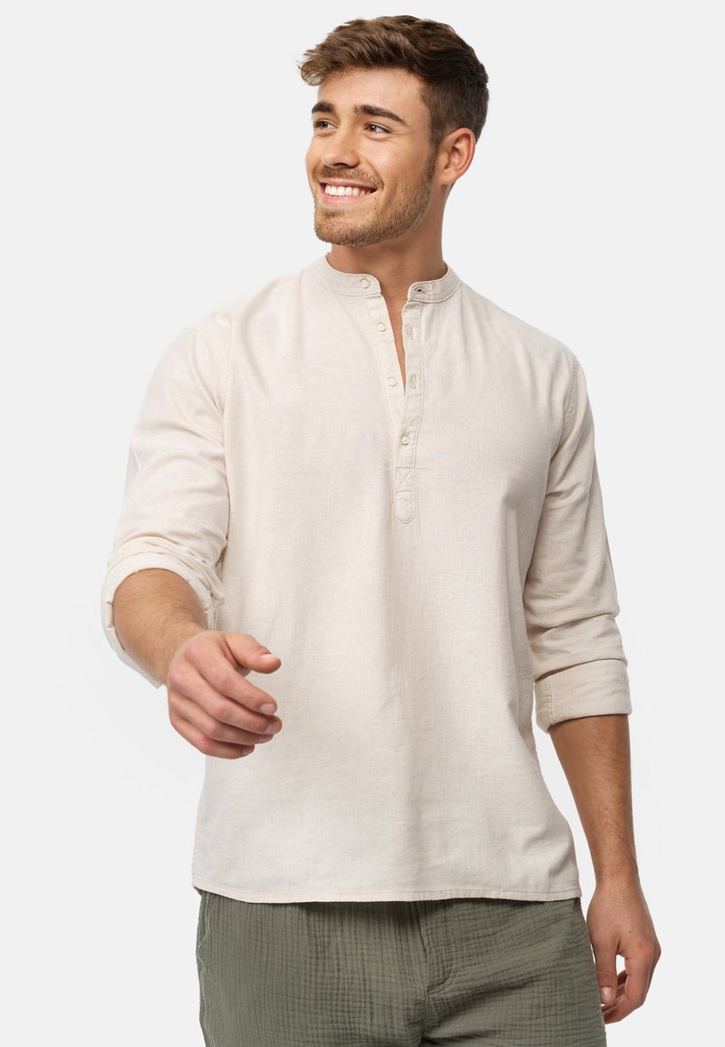 Camicia beige a maniche lunghe con colletto alla mandarino, patta frontale e maniche arrotolate, abbinata a pantaloni verde oliva con texture.