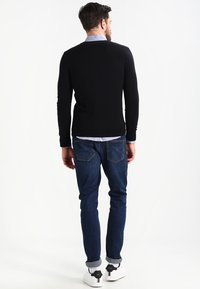 Svart tröja, blå skjorta med krage under, blå jeans och vita sneakers. Tröjan har en ren, välsittande design och ribbade ärmslut.