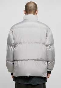 Puffjakke i lysegrå satinfinish med høj krave, horisontale baflestikninger og en løs, oversized silhuet.