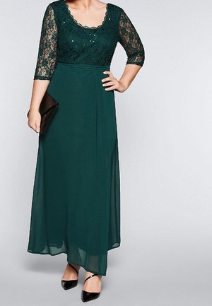 Vrouw draagt een donkergroen kanten topje en een chiffon rok, houdt een zwarte clutch vast, met zwarte pumps met spitse neus.