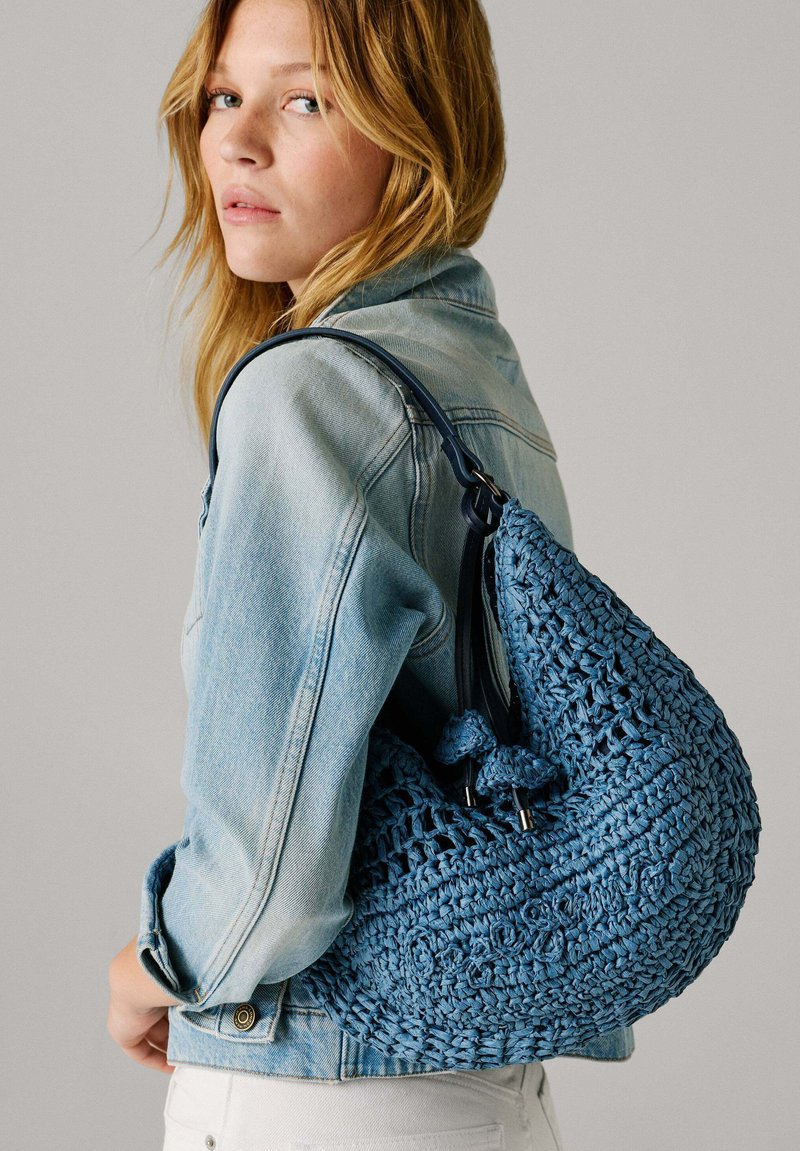Mlada ženska, ki nosi svetlo denim jakno in ima modro ramensko torbo z teksturo ter nastavljivimi trakovi.