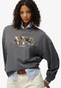 Sudadera gris con bordado floral y texto "Superdry", superpuesta a una camisa de rayas azul y blanca, combinada con pantalones de color claro.