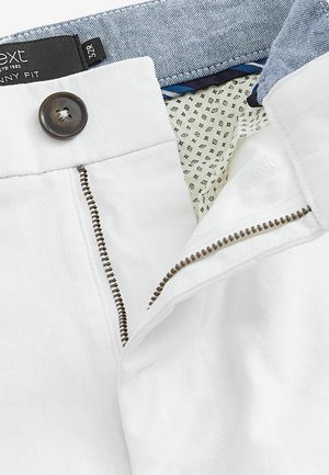 White skinny fit trousers with a zipper fly and brown button. Inside features a blue patterned waistband and a contrasting lined pocket.