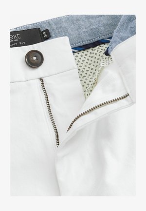 Weiße, enganliegende Hose mit Reißverschluss und braunem Knopf. Innen sind sie mit einem blauen gemusterten Bund und einer kontrastierenden gefütterten Tasche ausgestattet.