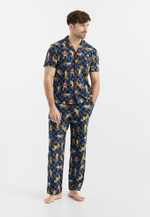 Mand iført marineblå ternet kortærmet skjorte og bukser med gule illustrationer af gepard og astronaut, stående barfodet.