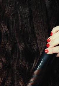 Prostownica do włosów o czarnym kolorze i eleganckim designie, z czarną, teksturowaną rączką, trzymająca ciemne, falowane włosy. Na dłoni widoczne są czerwone paznokcie.