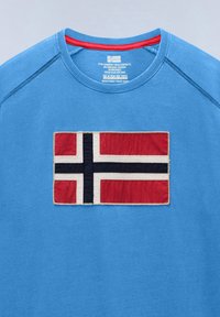 T-shirt bleu à col rond avec un patch cousu du drapeau norvégien centré sur la poitrine, avec des couleurs rouge, blanc et noir.