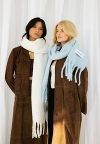 Deux femmes portent de longues écharpes : l'une blanche et l'autre bleu clair, toutes deux avec des franges. Elles sont toutes les deux associées à des vêtements extérieurs en daim marron.