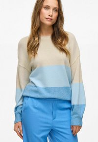 Sweter dzianinowy w beżowe i jasnoniebieskie poziome paski, z prążkowanymi mankietami i luźnym krojem, w zestawieniu z jasnoniebieskimi dopasowanymi spodniami.