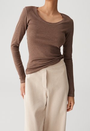 Mujer con una camiseta marrón ajustada de manga larga metida dentro de unos pantalones de talle alto, anchos y acanalados de color crema, frente a un fondo sencillo.