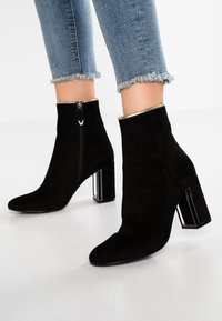 Black suede ankle boots with a pointed toe, silver zipper, and transparent block heel. Frayed denim jeans partially visible above the boots.