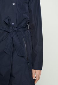 Marintrenchcoat med bälte i midjan, med tryckknappar och en ficka med dragkedja. Tillverkad av slät, vattenavvisande tyg med långa ärmar.