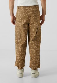 Pantaloni cargo maro cu un model repetitiv de logo în nuanță de bej, cu manșete elastice și multiple buzunare laterale. Asortați cu adidași albi.