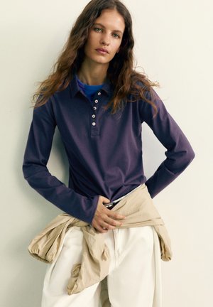 Une femme pose devant un mur clair. Elle porte une chemise bleu marine à manches longues, un pantalon blanc et une veste beige nouée autour de la taille.