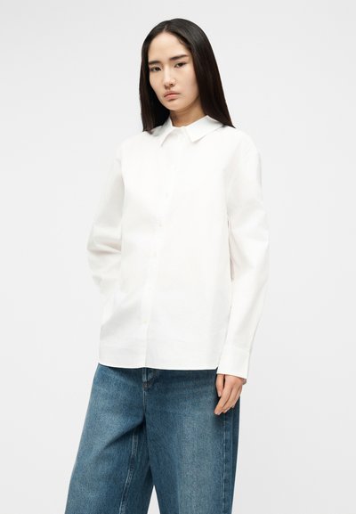 Ung kvinne iført en enkel hvit skjorte med knapper og vide blå jeans, stående mot en nøytral grå bakgrunn.