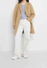 Abrigo de mezcla de lana de camello sobre un suéter de punto azul, combinado con jeans de pierna ancha blancos y zapatillas blancas. Líneas limpias y tonos neutros.