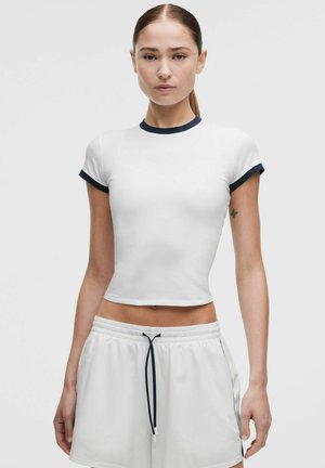 Žena s účesem staženým dozadu, která má na sobě přiléhavý bílý crop top s tmavým lemováním a volné bílé šortky s černým stahovacím páskem.
