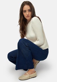 Suéter acanalado color marfil, jeans de campana azul oscuro y bailarinas de corte en color beige. La modelo está agachada con el cabello largo y ondulado.