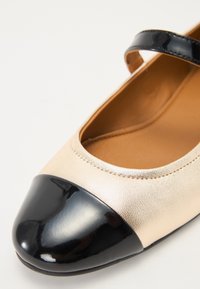Zapato de piel sintética beige con una puntera y una correa en cuero charol negro. Textura suave con un diseño redondeado y detalles de costura.