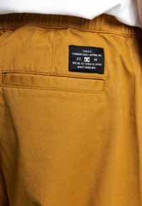 Hnědé šortky z tkané látky s černou nášivkou s informacemi o značce a specifikacemi, s zadní kapsou.