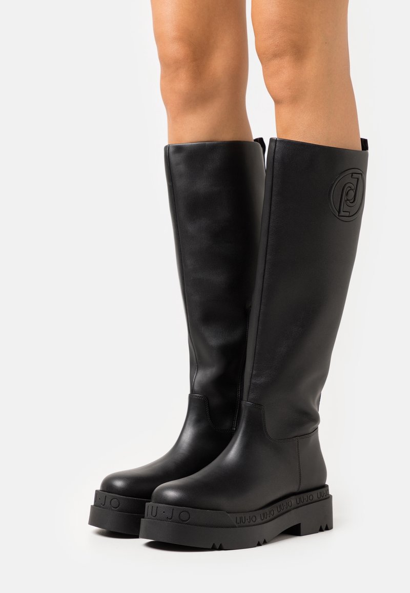 Bottes en cuir noires montant jusqu'aux genoux avec des semelles épaisses et un logo embossé sur la partie extérieure du mollet et la semelle, portées sur jambes nues sur un fond uni.