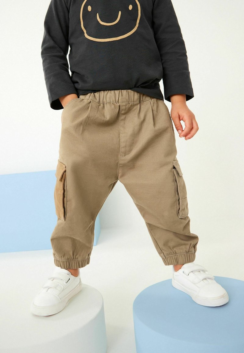 Pantalons cargo beiges avec des poignets élastiques et des poches latérales, associés à une chemise à manches longues noire ornée d'un graphique de visage souriant. Baskets blanches.