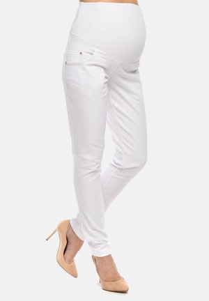 Pregnant woman wearing white maternity pants and beige high heels, standing with legs crossed against a plain background.