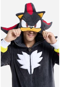 Tuta in peluche nera con cappuccio, caratterizzata da un design di personaggio; accenti rossi e gialli; zipper frontale; materiale in morbido pile; emblema ricamato bianco.