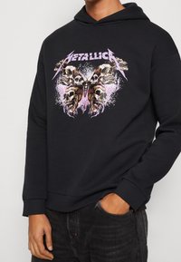 Černá mikina s kapucí, která má grafický design lebek a motýla, s fialovými akcenty a efekty stříkání. Mandlové rukávy a lem.