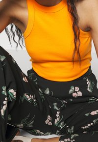 Pomarańczowy prążkowany top na ramiączkach w połączeniu z czarnymi spodniami w szerokie nogawki z kwiatowym wzorem w odcieniach różu i zieleni.