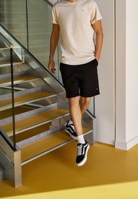 Beige bavlněné tričko, černé šortky a černé tenisky s bílými podrážkami. Stojí na kovových schodech s žlutými stupněmi a hladkou podlahou.