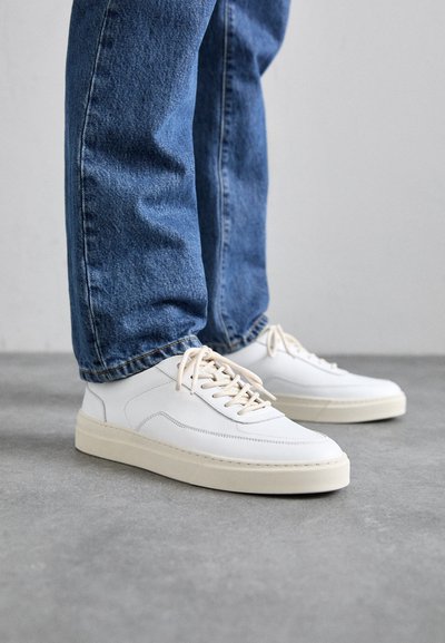 Weiße Leder-Sneaker mit cremefarbenen Gummisohlen, seitlichem Stich und beigen Schnürsenkeln. Kombiniert mit blauen Denim-Jeans.