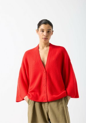 Red oversized cardigan with a deep V-neck, textured fabric, and five front buttons. Worn over beige wide-leg pants.