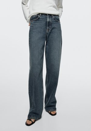 Persona che indossa jeans blu scuro larghi, maglione bianco e sandali neri con punta aperta, in piedi davanti a uno sfondo chiaro uniforme.