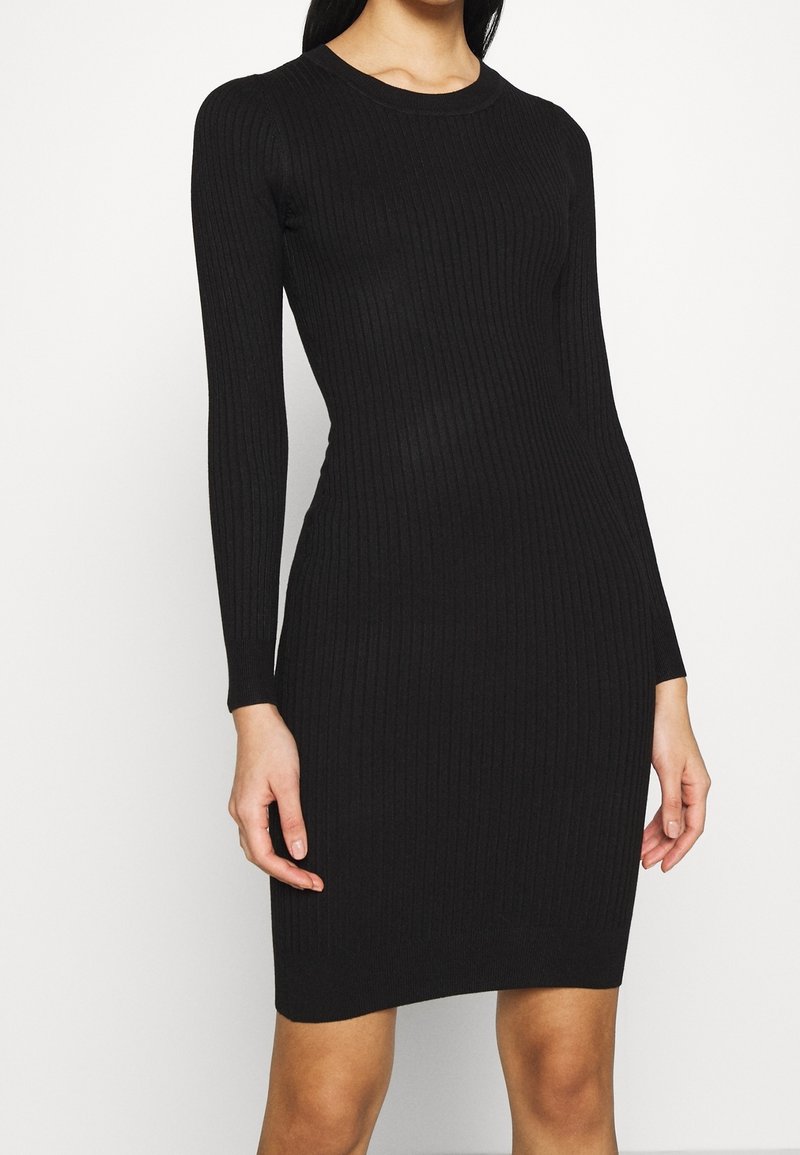 Femme portant une robe noire côtelée, ajustée et à manches longues, debout devant un fond clair uni.