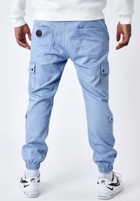 Uomo che indossa pantaloni cargo blu chiaro con polsini elasticizzati, scarpe Nike Air bianche e una maglietta bianca a maniche lunghe, mostrato di spalle.