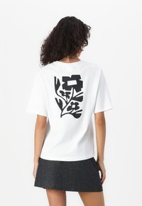 T-shirt en coton blanc avec un motif floral noir au dos. Manches courtes et coupe décontractée. Porté avec une jupe grise foncé.