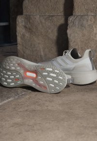 Coppia di scarpe sportive bianche con suole testurizzate con accenti bianchi e arancioni, posate su terreno di pietra contro un muro di pietra.